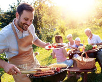 Man staat achter barbecue worstjes te grillen terwijl het gezin buiten aan tafel geniet van het eten